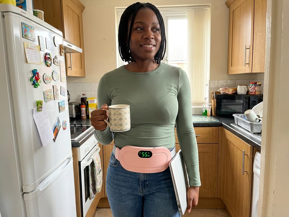 A woman standing in her kitchen with a mug and a laptop, wearing the heating belt